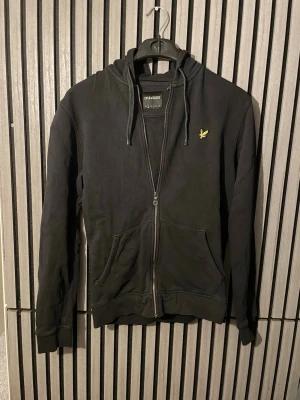 Svart hoodie med dragkedja - Svart hoodie från Lyle & Scott med dragkedja framtill, huva med dragsko och två fickor. Klassisk gul logga på bröstet. Perfekt för en avslappnad och stilren look.