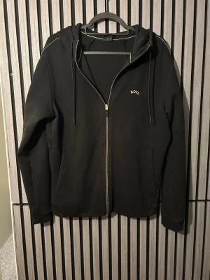 Svart hoodie med dragkedja från BOSS - Snygg svart hoodie från BOSS med dragkedja framtill och diskret logga på bröstet. Hoodien har huva med dragsko och vita kontrastsömmar på huvan och axlarna. Tillverkad i mjukt material och har två fickor framtill. Använd en del men riktigt bra skick. Pris kan diskuteras!