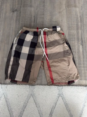 Burberry shorts med snöre  - Säljer ett par rutiga shorts i beige, svart, vitt och rött med bred resårmidja och vit snörning framtill. Shortsen har en loose passform och är perfekta för sommaren. Materialet känns lätt och syntetiskt, vilket gör dem sköna att bära varma dagar.