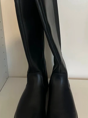 Svarta höga stövlar i skinnimitation - Snygga svarta knähöga stövlar i skinnimitation med elastisk panel längs skaftet för skön passform. Klassisk rund tå och låg sula gör dem perfekta till många olika outfits. Stövlarna har en stilren och minimalistisk design.