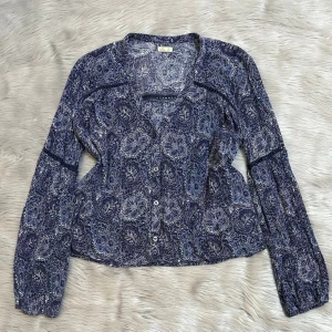 Hollister Blå blus med blommigt mönster - Säljer en blå blus med vitt och blått blommigt mönster. Blusen har långa ärmar, knappar framtill och dekorativa sömmar vid axlar och ärmar. Tunn och luftig, perfekt för lager på lager eller till jeans.