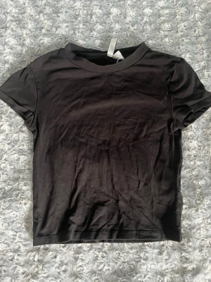 Svart basic t-shirt i bomull - En enkel svart t-shirt i mjuk bomull med rund hals och korta ärmar. Perfekt till jeans eller shorts för en clean och avslappnad look. Passformen är normal och tyget känns skönt mot huden.