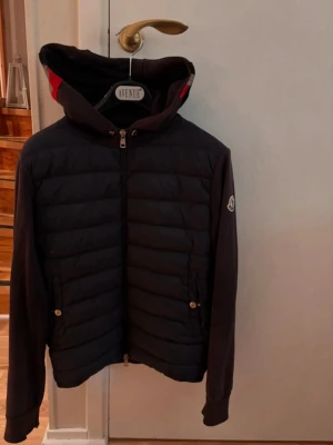 Moncler cardigan - Svart cardiganjacka från Moncler med vadderad framsida och mjuka ärmar. Jackan har huva med röd och grå rand, dragkedja framtill och två fickor med knapp. Moncler-logga på ärmen ger en exklusiv touch. Perfekt för kyliga dagar.