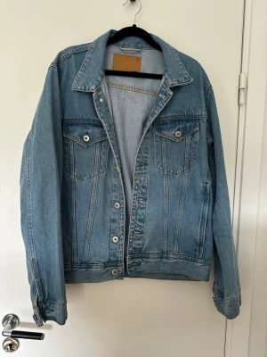 Klassisk blå jeansjacka unisex M - Snygg blå jeansjacka i klassisk modell med raka sömmar och två bröstfickor med lock och knapp. Jackan har silverfärgade knappar och justerbara ärmslut. Perfekt att slänga över en hoodie eller t-shirt för en avslappnad look. 