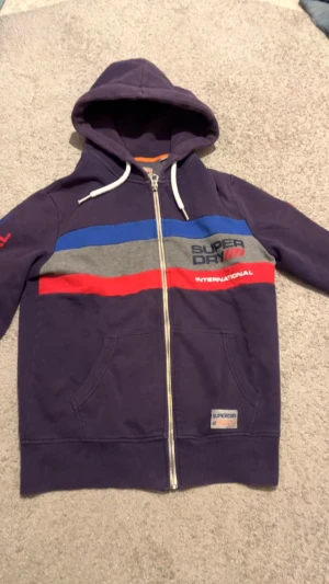Lila Superdry hoodie med dragkedja - Snygg lila hoodie från Superdry med dragkedja, huva och vita dragsnören. Framsidan har tryck i blått, grått och rött med texten 'Superdry International'. Praktiska fickor framtill och ribbade muddar. Perfekt för en avslappnad streetstyle.