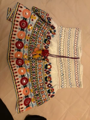 Broderad kjol med speglar och tofsar - Färgglad kortkjol med broderade blommor i rött, orange, gult och blått. Kjolen har speglar, tofsar och detaljer i flera färger samt snörning i midjan. Materialet känns som bomull och kjolen har en unik, handgjord känsla med volangkant.