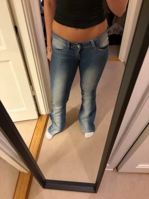 Ljusblå bootcut jeans med låg midja - Säljer ett par ljusblå bootcut jeans med låg midja och snygga slitningar framtill. Jeansen har klassisk femficksdesign och är tillverkade i stretchigt denimtyg för skön passform. Perfekta för dig som gillar retrovibbar och en avslappnad look.