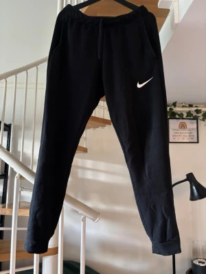 Svarta mjukisbyxor från Nike - Snygga svarta mjukisbyxor från Nike med vit logga på vänster ben. Byxorna har dragsko i midjan, muddar vid bensluten och två sidofickor. Perfekta för chill eller träning, tillverkade i mjukt material för maximal komfort. Byxorna har ett jättelitet brännmärke på vänster ben