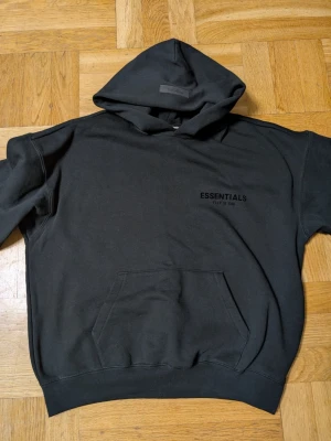 Black Essentials Fear of God hoodie - Säljer en Black hoodie från Essentials Fear of God med stor huva och känguruficka. Tryck med 'ESSENTIALS' på bröstet och ryggen. Oversized passform och mjukt material som känns riktigt skönt. Perfekt för dig som gillar streetwear och enkel stil. Har aldrig använts pga storleken inte passa mig. Skicka in era bud så kanske accepteras dem