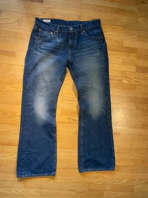 Blå Levis 527 bootcut - Säljer ett par klassiska Levis 527 blå jeans med bootcut passform och snyggt slitna detaljer på låren. Jeansen har fem fickor, gylf och kontrastsömmar. Materialet är kraftig denim i bomull. Perfekta för en avslappnad och tidlös stil.