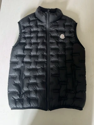 Svart dunväst från Moncler - Snygg svart dunväst från Moncler med quiltat mönster och hög krage. Västen har två dragkedjeförsedda fickor och en klassisk Moncler-logga på bröstet. Perfekt för lager-på-lager och håller dig varm med stil.