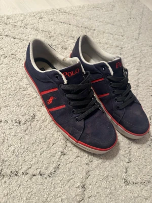 Marinblå Polo Ralph Lauren sneakers - Snygga marinblå sneakers från Polo Ralph Lauren med röda detaljer och logga på sidan. Skorna har mörkblåa snören, vit sula med röd rand och är tillverkade i canvasmaterial. Perfekta för en avslappnad och trendig stil. Storlek 43, original boxen har jag inte kvar men kommer i en annan skokartong! Hör av er vid minsta fundering och kolla gärna in mina andra annonser!