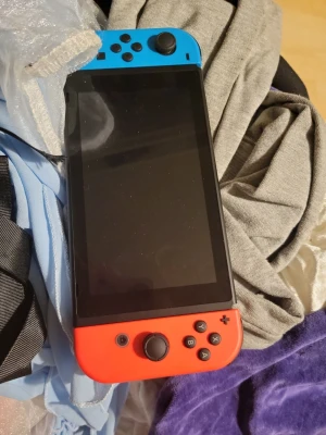 Nintendo Switch med blå och röd Joy-Con + tomodatchi life - Nintendo Switch spelkonsol med en stor skärm och avtagbara Joy-Con-kontroller i blått och rött. Konsolen har en rektangulär form och är tillverkad i plast. Perfekt för gaming både hemma och på språng.
