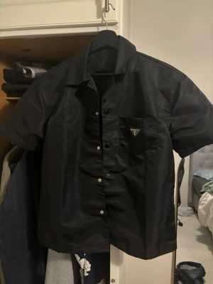 Prada tröja - Snygg svart skjorta från Prada med korta ärmar och klassisk krage. Skjortan har knappar framtill och en bröstficka med det ikoniska Prada-emblemet i metall. Tillverkad i ett glansigt syntetmaterial som ger en modern look.