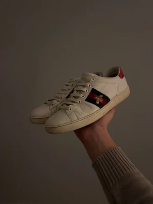 Vita sneakers med broderad bi - Snygga vita sneakers med röd och grön sidodetalj samt en broderad guldfärgad bi på sidan. Skorna har lågt skaft, vita snören och röd detalj på hälen. Klassisk och stilren design i läder med gummisula.