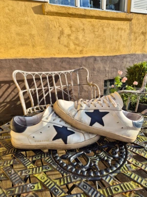 Vita golden goose sneakers med blå stjärna - Snygga vita golden goose sneakers med mörkblå stjärna på sidan och matchande blå detalj vid hälen. Skorna har en klassisk låg siluett och är tillverkade i läder med beige sula. Perfekta för en avslappnad och trendig stil.