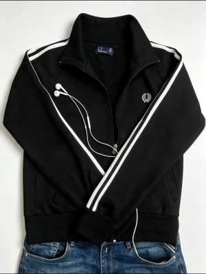 Svart track jacket från Fred Perry - Snygg svart track jacket från Fred Perry med vita ränder längs ärmarna och klassisk logga på bröstet. Dragkedja framtill, hög krage och ribbade muddar. Perfekt till jeans eller träningsbyxor för en sportig look.