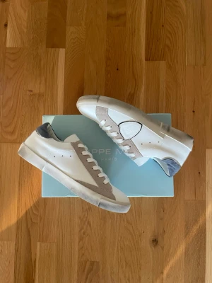 Vita och grå sneakers från Philippe Model - Snygga vita sneakers från Phillipe Model med grå mockadetaljer och vit sula. Skorna har en unik design med en sköldformad detalj på sidan och grå hälparti. Perfekta för dig som gillar stilrena och trendiga sneakers med en twist.