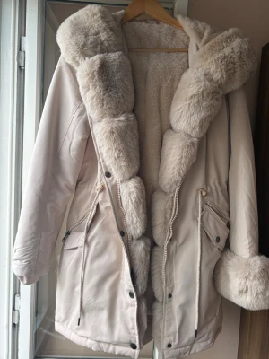 Beige parkas med fuskpäls och snörning - Snygg beige parkas med fluffig fuskpäls runt huva, ärmslut och insida. Jackan har dragkedja, knappar, snörning i midjan och stora fickor med dragkedja och lock. Perfekt för kalla dagar och ger en lyxig känsla.