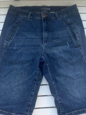 Blå denimshorts med fickor - Säljer ett par klassiska blå denimshorts med normal passform. Shortsen har framfickor, bälteshällor och knappstängning. Baksidan har två dekorativa fickor. Perfekta för sommardagar och enkel att matcha med olika stilar.