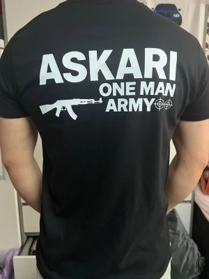 Svart T-shirt – Askari One Man Army - Svart T-shirt med “Askari One Man Army”-tryck på fram- och baksidan. I mycket fint skick, använd endast ett fåtal gånger. Bekväm passform och perfekt för vardagsbruk.
