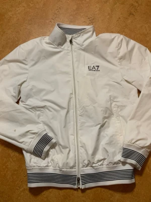 Vit vindjacka från EA7 Emporio Armani - Snygg vit vindjacka från EA7 Emporio Armani med dragkedja framtill och ribbade muddar med svarta ränder. Jackan har hög krage och två sidofickor. Perfekt för en sportig och stilren look.