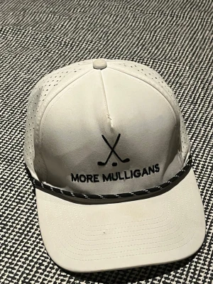 Vit keps med text More Mulligans - Snygg vit keps med svart texten 'MORE MULLIGANS' och två korsade golfklubbor framtill. Kepsen har perforerade sidor för extra ventilation och ett svartvitt snöre längs skärmen. Justerbar baktill och tillverkad i lätt material, perfekt för golf eller vardagsstil.