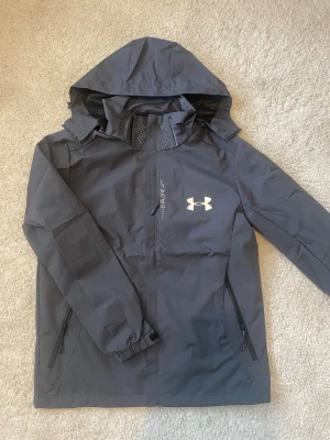Under armour vind/regn jacka - mörkgrå - Snygg oanvänd under armour jacka. Har 2 stora fickor på framsidan och 1 vid bröstet. Passar som en stor S och liten M. 
