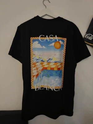Svart Casa Blanca t-shirt med tryck - Säljer en svart t-shirt från Casa Blanca med färgglatt tryck av pool och sol på både fram- och baksida. T-shirten har rund hals och korta ärmar. Perfekt för dig som gillar grafiska motiv och vill sticka ut lite extra.