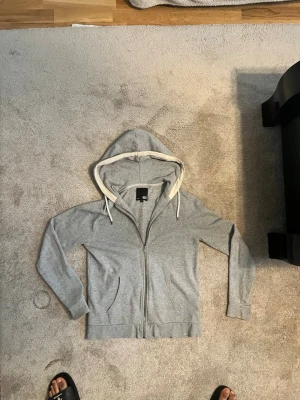 Ljusgrå zip hoodie med huva - Ljusgrå hoodie med dragkedja från 157. Tröjan har en stor huva med vit dragsko och kontrasterande ljus insida. Klassisk känguruficka framtill och ribbade muddar vid ärmar och nederkant. Perfekt för en chill och avslappnad stil. Strlk M