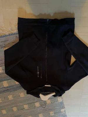 Svart hoodie från Sail Racing - Svart hoodie från Sail Racing med dragkedja framtill och diskret logga på bröstet. Klassisk modell med huva och mjukt material på insidan. Perfekt för dig som gillar stilrena och sportiga plagg. Passar till det mesta och är riktigt bekväm.
