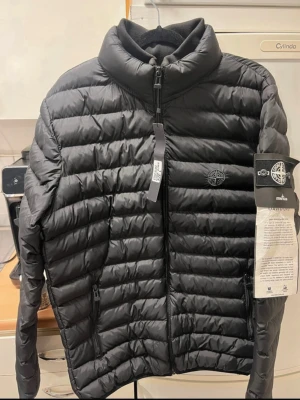 Svart dunjacka från Stone Island - Snygg svart dunjacka från Stone Island med quiltad design och hög krage. Jackan har dragkedja framtill, två sidofickor med dragkedja och den klassiska Stone Island-loggan på bröstet. Perfekt för kalla vinterdagar och riktigt stilren look.