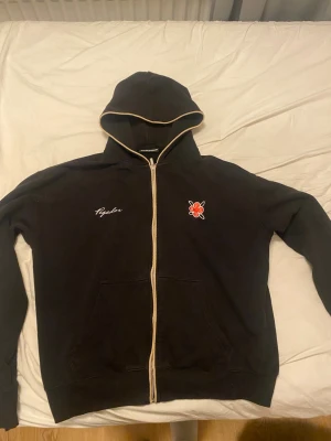 Svart hoodie med dragkedja och brodyr - Svart hoodie med vit dragkedja och kontrastkant på huvan från Pegador. Broderad text och röd logga på bröstet. Mjuk bomullskänsla, två fickor fram och klassisk passform. Perfekt för en chill och stilren look. Hör gärna av er vid frågor!