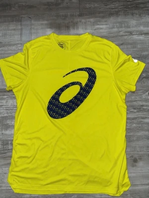 Gul tränings-tshirt från Asics - Snygg gul tränings-tshirt från Asics med stort svart logotryck på bröstet. Tillverkad i ett lätt och snabbtorkande funktionsmaterial som passar perfekt till löpning eller gym. Klassisk rund hals och korta ärmar.