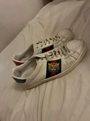 Vita sneakers med tiger och ränder - Snygga vita sneakers med gröna och röda ränder på sidan samt en tigerpatch. Skorna har lågt skaft och detaljer i grönt och rött på hälen. Tillverkade i läder med klassisk snörning och vit sula. Perfekta för dig som vill sticka ut med coola detaljer.