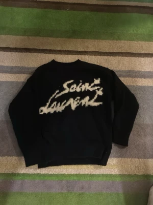 Svart stickad Saint Laurent-tröja - Svart stickad tröja från Saint Laurent med stort vitt logotyptryck i graffiti-stil på framsidan. Tröjan har rund halsringning och långa ärmar. Perfekt för dig som vill ha en statement-piece i garderoben.