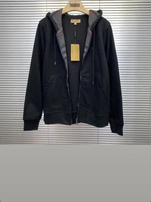 Svart hoodie från Burberry - Snygg svart hoodie från Burberry med dragkedja och klassiskt rutigt foder i huvan. Hoodien har två fickor fram och en diskret broderad logga på bröstet. Perfekt för dig som gillar stilrena plagg med exklusiv känsla.
