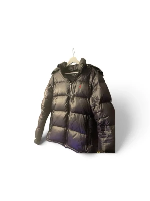 Svart dunjacka med huva - Perfekt inför vintern!Snygg svart dunjacka med huva och dragkedja framtill. Jackan har en puffig design med breda kanaler och en liten röd logga på bröstet. Perfekt för kalla dagar och riktigt skön att bära. Passar dig som vill ha en stilren och varm jacka.