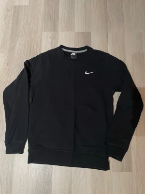 Svart sweatshirt från Nike - Klassisk svart sweatshirt från Nike med rund halsringning och vit broderad logga på bröstet. Tröjan har långa ärmar och ribbade muddar vid ärmslut och nederkant. Perfekt för en sportig och avslappnad stil.