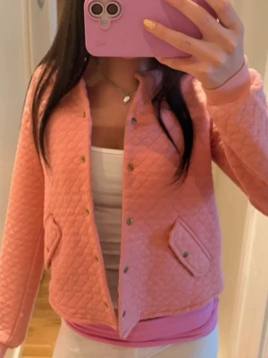 Quiltad rosa cardiganjacka - Säljer en quiltad rosa cardiganjacka med tryckknappar i guld och två fickor framtill. Jackan har en mjuk, vadderad känsla och ribbade muddar vid ärmslut och nederkant. Perfekt för dig som vill ha en söt och trendig look.
