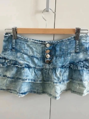 Blå volangkjol i denim - Säljer en kort blå jeanskjol med volanger och dekorativa knappar framtill. Kjolen har bälteshällor och en tvättad denimlook som ger en avslappnad vibe. 