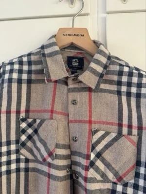 En burberry skjorta  - Stilren rutig skjorta i beige med blå, vita och röda ränder. Skjortan har två bröstfickor med knappstängning och klassisk krage. Perfekt för en avslappnad men trendig look.