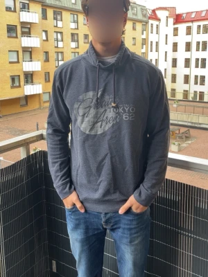 Vintage Tom tailor tröja - Säljer en mörkgrå hoodie med tryck på bröstet där det står 'Tokyo Area 62'. Perfekt för en avslappnad och stilren look till vardags. Hoodie har ingen luva, istället en krage, som ni ser på bild 4. Storlek L, modellen är 190cm!