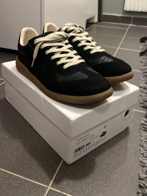 Masion martin marigela - Stilrena svarta sneakers med beige gummisula och vita snören. Ovandelen är i mocka och läder, vilket ger en snygg kontrast och modern look. Perfekta för dig som gillar klassisk men trendig stil.