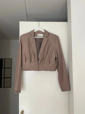 Kort beige kavaj med en knapp - Snygg beige kavaj i kort modell med klassisk krage och en knapp framtill. Kavajen har långa ärmar och är fodrad på insidan. Perfekt för en trendig och stilren look.