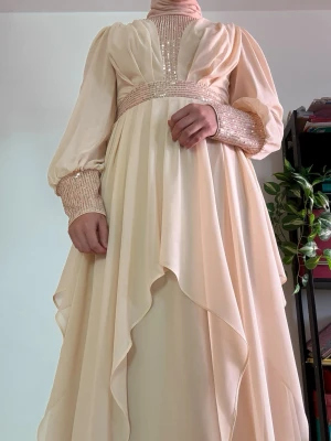 Ljusbeige långklänning med volang - Elegant långklänning i ljusbeige med volangdetaljer och smockade muddar. Klänningen har långärmade puffärmar och hög hals med dekorativa paljetter i midja och överdel. Flödande material ger en drömmig look.