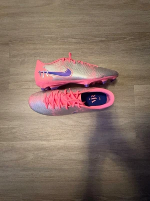 Nike rosa och silver fotbollsskor - Säljer ett par Nike fotbollsskor i en snygg rosa och silver färg med lila detaljer och ViniJR-logga på sidan. Skorna har snörning, låg profil och fasta dobbar för spel på gräs. Perfekta för dig som vill sticka ut på planen!