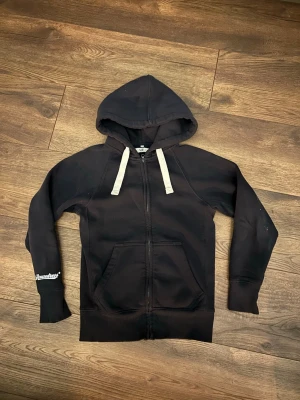 Mörkblå Russemerch - Mörkblå Russemerch hoodie med dragkedja och vita tjocka snören i huvan. Ficka framtillen men även inne till vänster och tryckt text på ena ärmen. Klassisk passform och mjukt material, perfekt för en avslappnad stil.