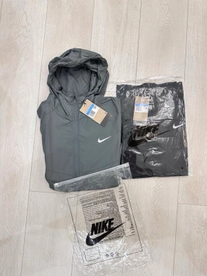 Grå vindjacka med huva från Nike - Snygg grå vindjacka från Nike med huva och dragkedja framtill. Jackan har en minimalistisk design med vit Nike-logga på bröstet och är tillverkad i lätt, vindtätt material. Perfekt för blåsiga dagar och enkel att matcha med olika outfits.