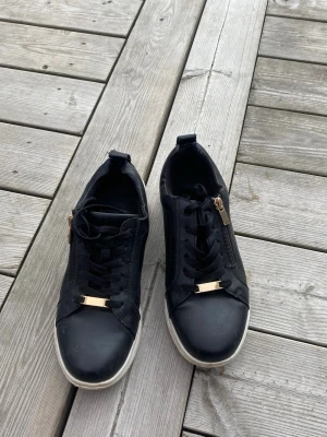 Svarta sneakers med gulddetaljer - Snygga svarta sneakers med vita sulor och guldfärgade detaljer på snörningen och dragkedjan på sidan. Skorna har en låg profil och är tillverkade i en mix av textil och syntetmaterial. Perfekta för en trendig och avslappnad look.